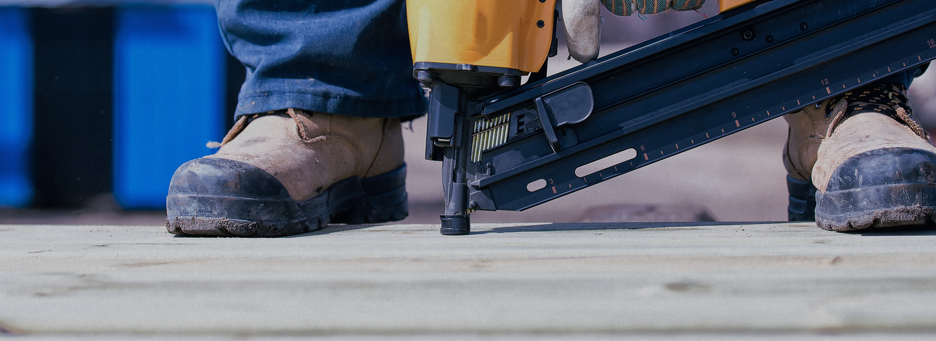 Worker's boots next to a nail gun on wooden planks, with a blue background.